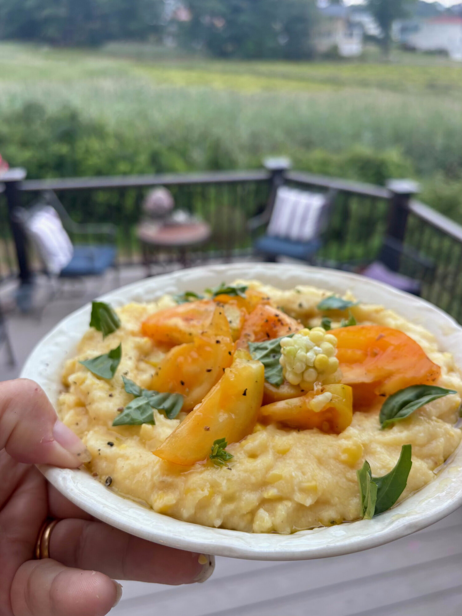 Bowl of corn polenta with tomatoes held up on a porch overlooking a salt marsh