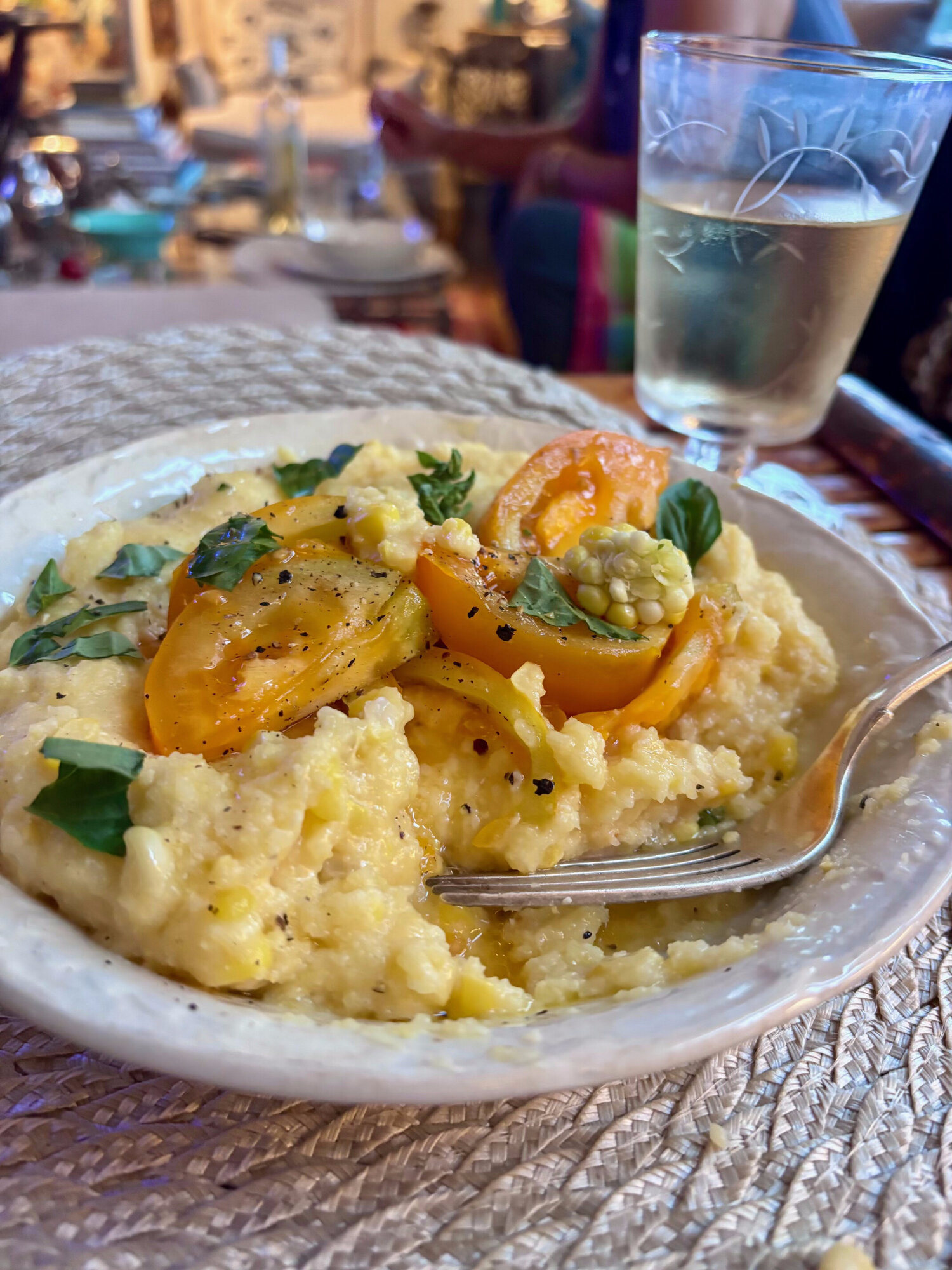 Bowl of creamy corn polenta topped with chopped tomatoes, basil and olive oil, with a fork and a glass of white wine