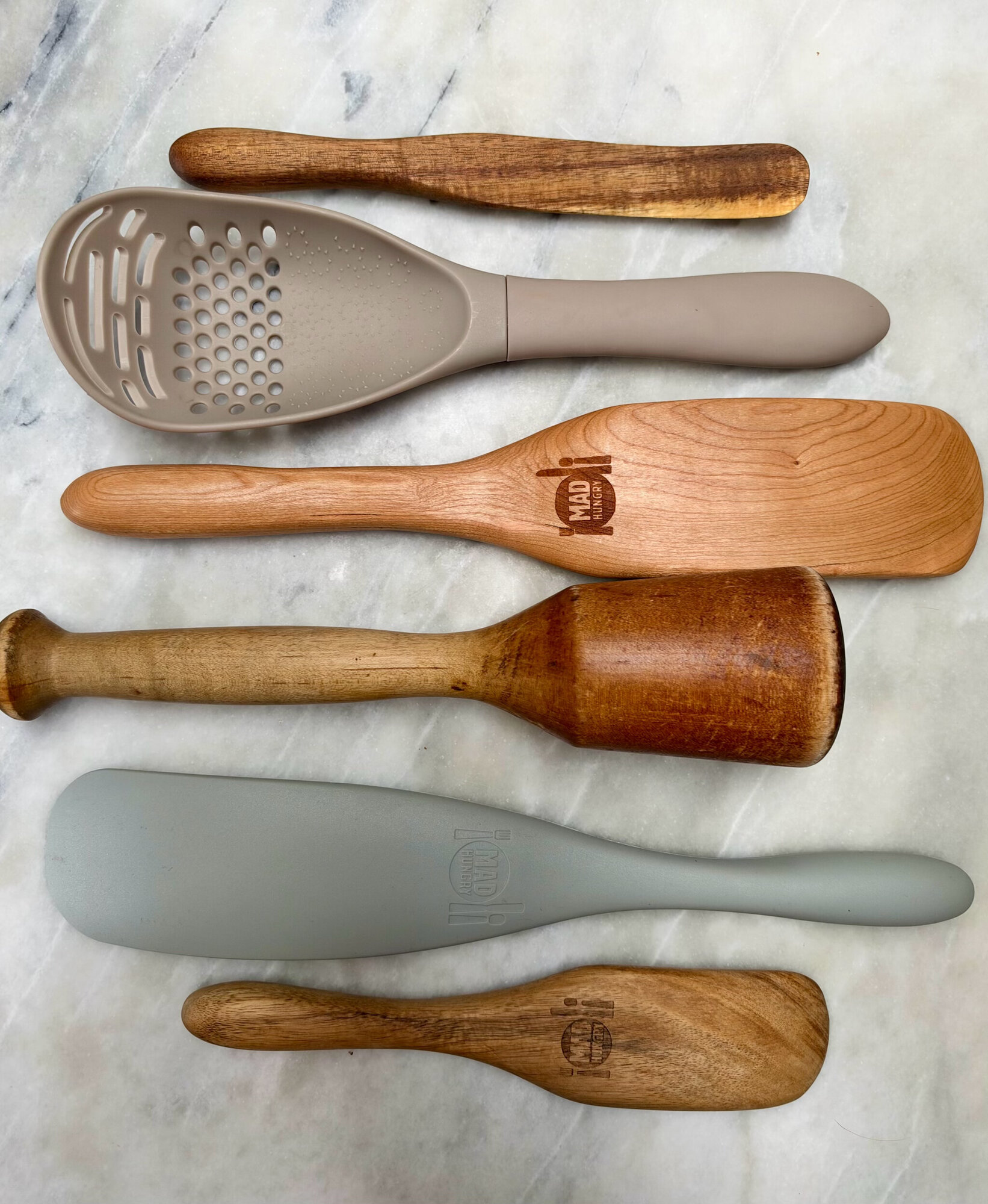 Six kitchen tools on marble: Mad Hungry acacia and cherry wood spurtles, a silicone Super Spoon, a silicone spurtle, and a vintage wooden Beetle
